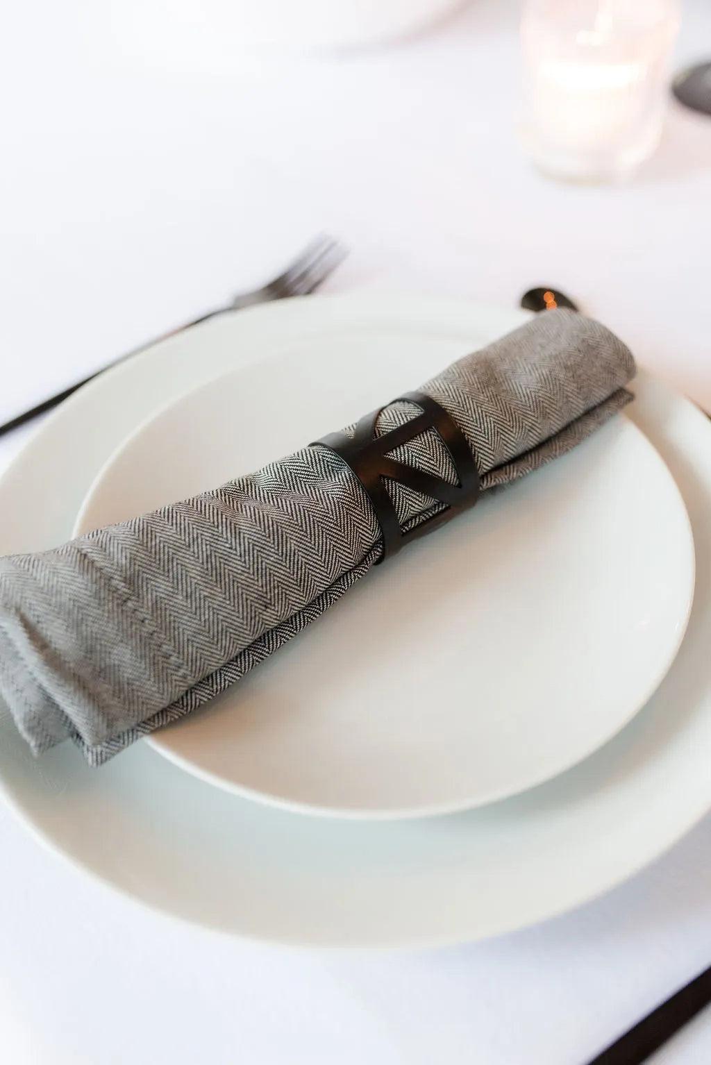 Black metal napkin ring holding a grey napkin sitting on two white plates with black cutlery on a white tablecloth
