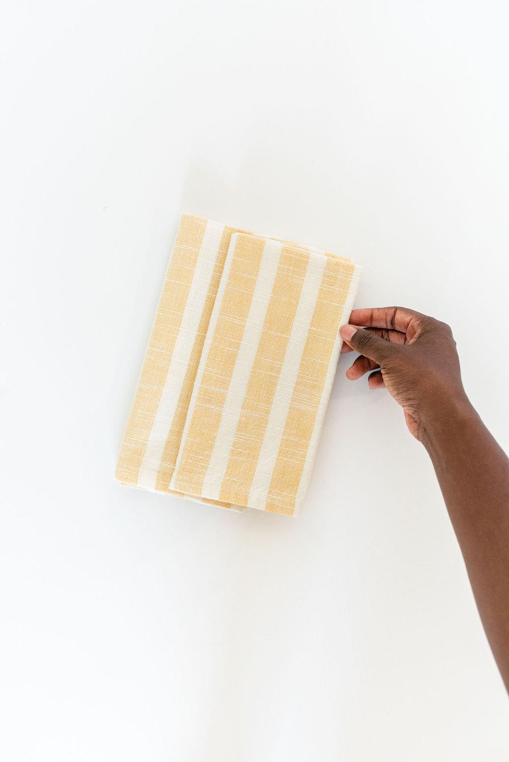 two folded yellow striped cotton napkins on an angle white background with hand holding bottom right corner of napkin