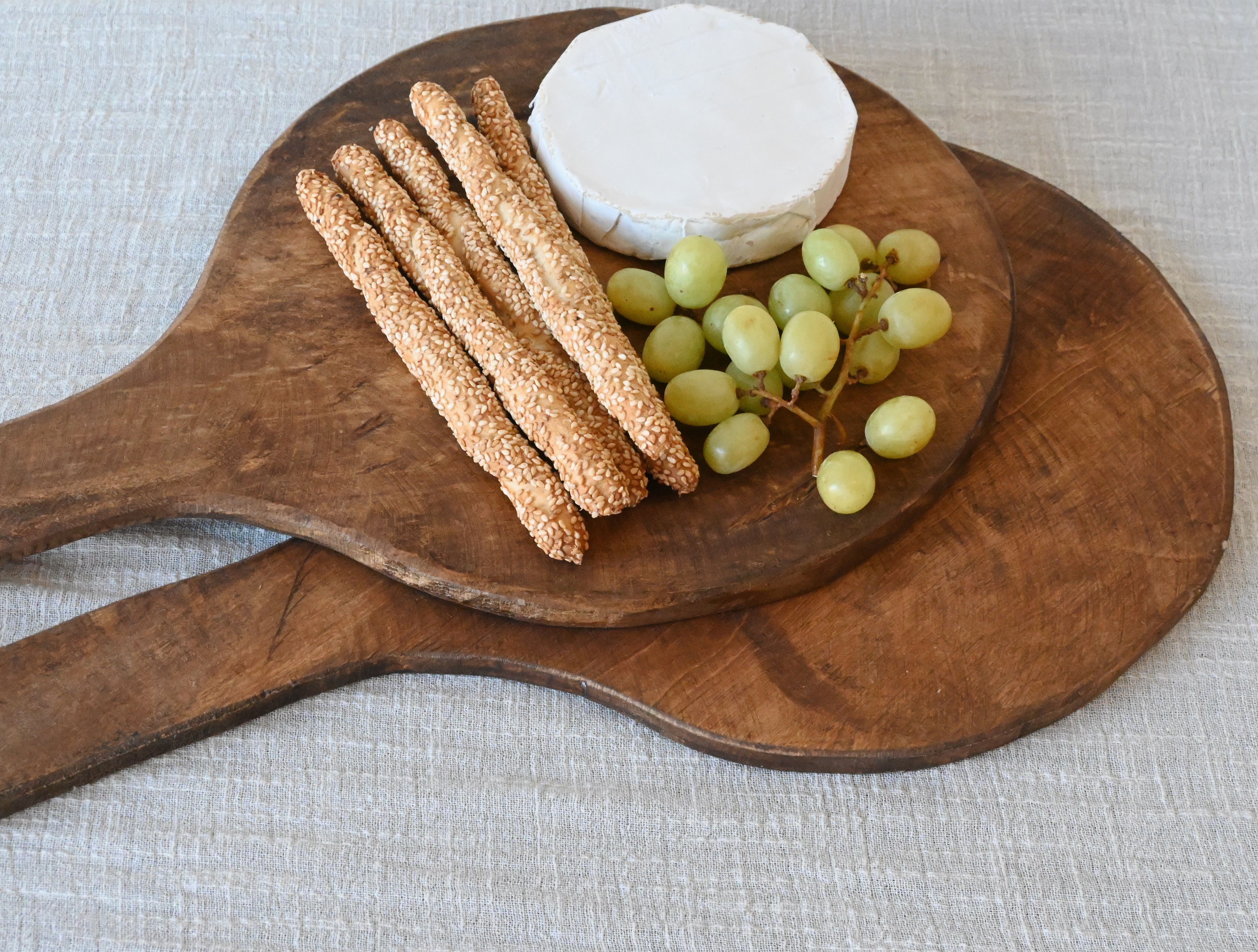 two round mango wood charcuterie or cutting boards sitting on top of one another. One board has bread sticks, brie and green grapes