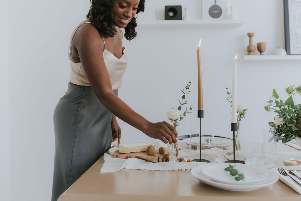 woman putting white rose in small glass vase next to two onoir tapered candle holders with candles on table with table runner