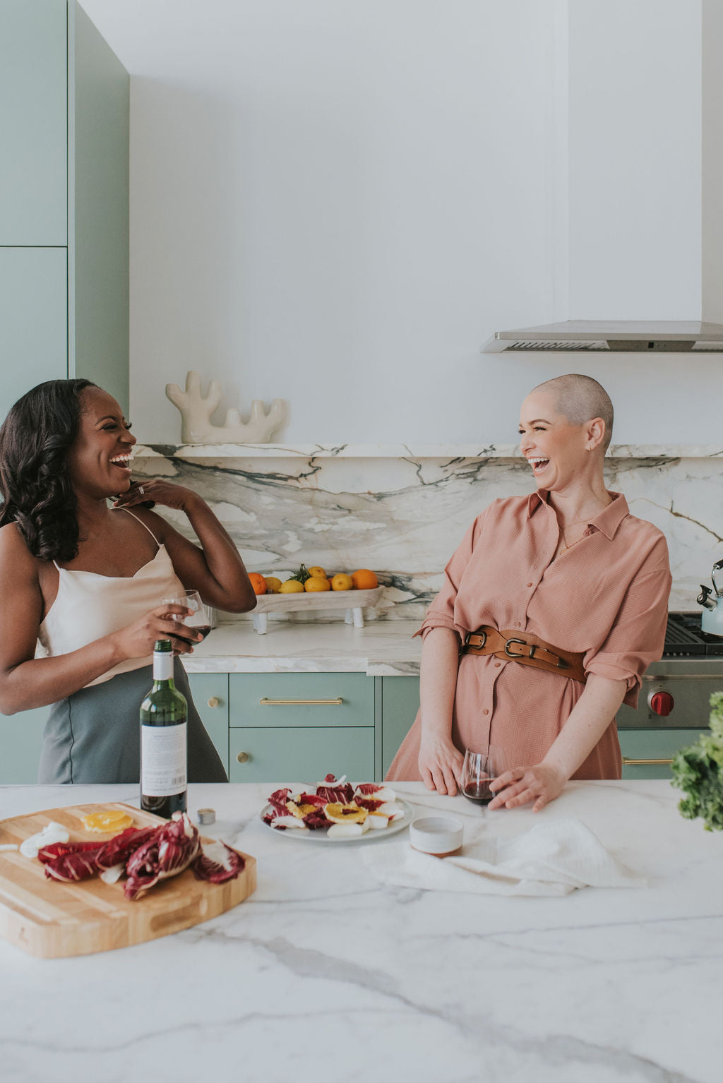 two beautiful smiling women in a kitchen with white marble counters, wine and a nice platting of citrus and radicchio
