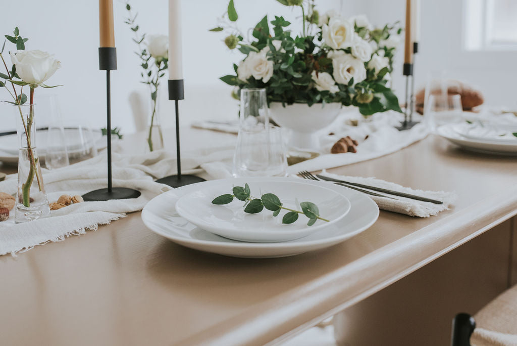 light wood table with white table runner, with 4 white plates table settings,and 4 onoir taper candle holders with candles