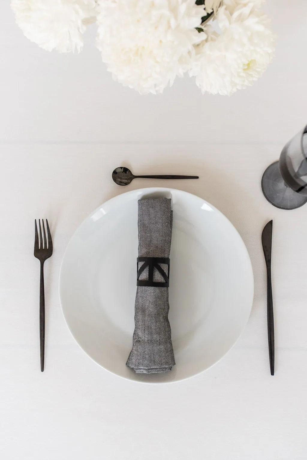 Black metal napkin ring holding a grey napkin sitting on a white plate with black cutlery on a white table cloth with a white flower centerpiece